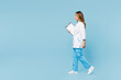 © ViDi Studio - Full body side view female doctor woman wear white gown suit work in hospital clinic office holding clipboard with medical documents go isolated on plain blue background. Health care medicine concept.