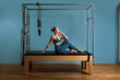 © Georgii - A young girl does Pilates exercises with a bed reformer, barrel machine tool. Beautiful slim fitness trainer on the background of a reformer doing various exercises gray, low key, light art. Fitness