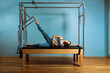 © Georgii - A young girl does Pilates exercises with a bed reformer, barrel machine tool. Beautiful slim fitness trainer on the background of a reformer doing various exercises gray, low key, light art. Fitness