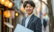 © Natalia - Young asian man in business suit holding laptop. Generative AI.