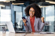 © Liubomir - Cheerful african american female in business suit with mobile gadget in hand making victory gesture in office. Rejoiced worker with toothy smile celebrating triumph by computer during daytime