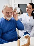Fototapeta Tulipany - Mature man with ITE hearing aid looks at himself in mirror held by smiling audiologist, and tries on hearing device. Stylish hearing device
