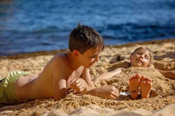 Canvas Print - Child, tickling sibling on the beach on the feet with feather, kid cover in sand, smiling, laughing