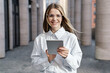 © muse studio - Young professional woman outdoors holding tablet, smiling confidently in urban setting.