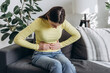 © Yura Yarema - Close up of upset young woman suffering from menstrual pain. Girl with hands squeezing belly having painful stomach ache or period cramps sitting on couch. Abdominal pain and painful periods concept