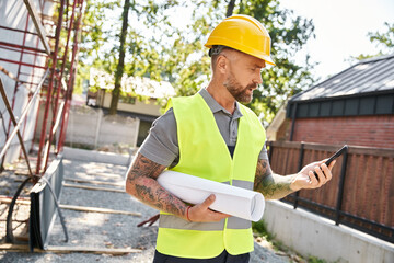 handsome bearded construction worker with tattoos and blueprint looking at phone, cottage builder