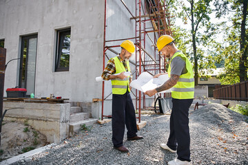 attractive bearded cottage builders in safety vests working on their blueprints on construction site