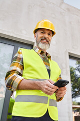 vertical shot of jolly man in safety vest posing with phone and looking at camera, cottage builder