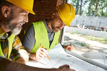 two handsome builders in safety helmets and vests working with blueprint before construction