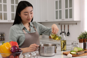 Cooking process. Beautiful woman pouring oil from bottle into pot at white countertop in kitchen