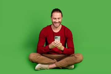 Wall Mural - Full length photo of satisfied man dressed pullover brown pants sit look at smartphone chatting isolated on green color background