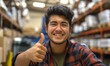 Young cheerful latino or hispanic man smiling at camera with toothy smile and his thumbs up at warehouse distribution packaging centre for logistics and delivery purposes