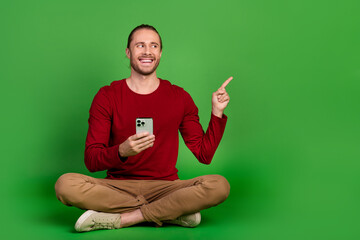 Full size photo optimistic guy wear red shirt sit with smartphone look directing at promo empty space isolated on green color background