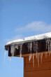 Many melting icicles with fall down drops hangs down close-up on the roof edge with snow against blue spring sky