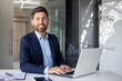 © Tetiana - Portrait of a successful young businessman in a suit sitting at a desk in the office, working with documents and using a laptop, confidently and smilingly looking at the camera