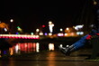 Nogi osoby ubranej w spodnie jeansowe siedzącej na ławce, w tle światła miasta nocą odbijające się w wodzie