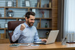 © Liubomir - Happy young business man sitting in the office at the desk with a notebook and rejoices at the success, looking at the window, showing a victory gesture with his hand