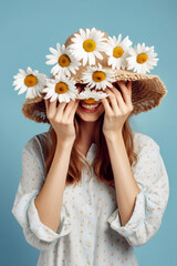 Portrait of a young cheerful woman with a hat decorated with a daisy flower wreath on her head and shyly covering her face with her hands, on a blue background.