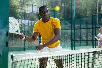 African-american man in t-shirt playing padel tennis on court. Racket sport training outdoors.