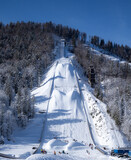 Fototapeta Tulipany - Giant Planica Ski Jump with Snow-Covered Slopes on a clear sunny winter day. People playing on snow at the bottom of the ski jump