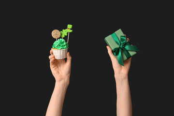 Female hands with tasty cupcake and gift box for St. Patrick's Day on black background