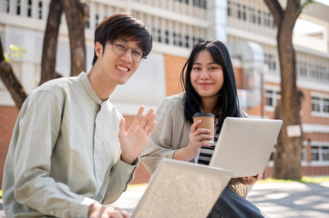 Wall Mural - Two smiling Asian students are sitting on a bench in the university park together with their laptop.