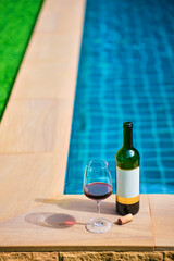 A bottle of red wine and a glass of wine are set on the edge of a clear blue pool, surrounded by a lush green lawn. The afternoon sun shines down, creating a relaxing and inviting atmosphere.