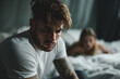 handsome model man sitting on the edge of his bed worried expression, in the background a woman looking at him. relationship problems