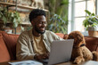 black handsome model man is working from home on his laptop, video call, smiling, accompanied by his dog. 