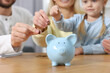 © New Africa - Planning budget together. Little girl with her family putting coins into piggybank at table, closeup
