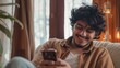 A happy curly young Indian man chats with girlfriend at home from a comfortable armchair, using a modern mobile phone to check social media and use a mobile app. There is copy space on the panorama.