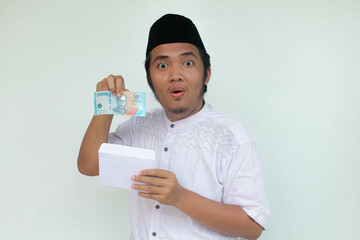 A young Asian muslim man holding an envelope and fifty thousand rupiah in cash with an expression of surprise and happiness. THR (Tunjangan Hari Raya) or Religious Holiday Allowance Concept.