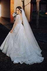 A brunette bride in a white dress with a long train holds the dress and walks down the stone path. Autumn. Wedding photo session in nature. Beautiful hair and makeup. celebration