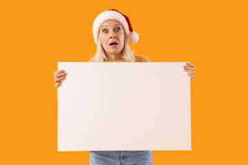 Sticker - Surprised mature woman in Santa hat holding blank poster on yellow background