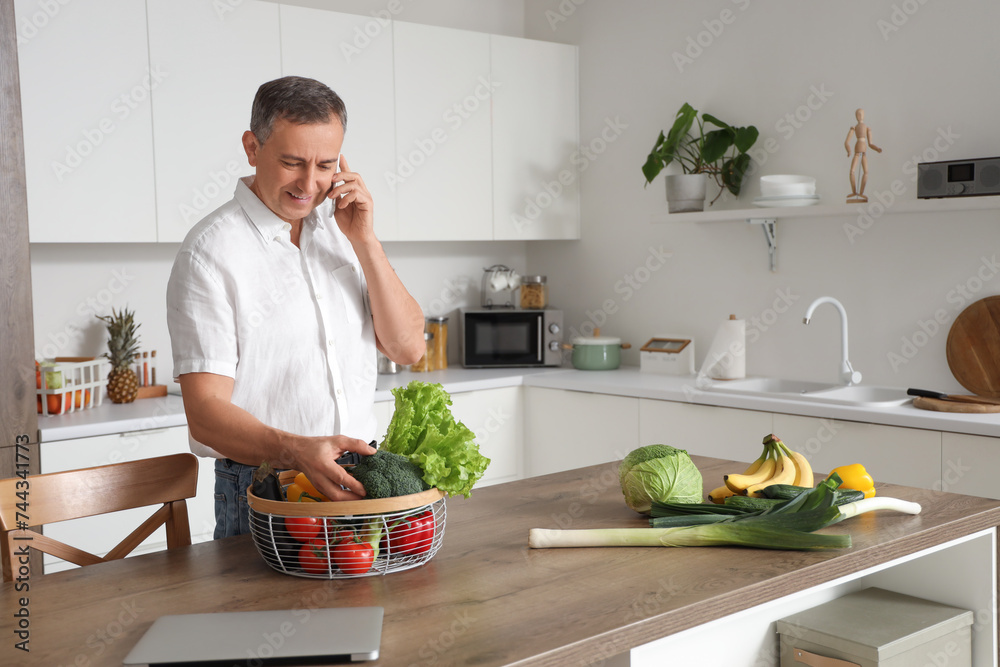 Mature man with fresh vegetables talking by mobile phone in kitchen