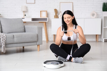 Poster - Beautiful young sporty woman with scales and measuring tape sitting at home. Weight loss concept