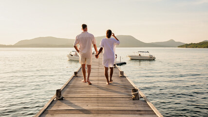 Wall Mural - A man and a woman standing on a dock by a lake, holding hands. They are wearing shorts and looking out at the horizon, happy and relaxed during their leisurely travel in Thailand, a couple wooden pier