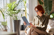 © insta_photos - Busy elegant young professional business woman executive with red hair holding digital tablet working sitting in comfortable chair in sunny office with green plants. Candid authentic phot, copy space