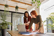 © insta_photos - Two smiling professional female partners or coworkers, happy business women entrepreneurs working together in office looking at laptop using computer writing notes standing at work desk.
