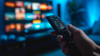 © HelenP - close-up view of a hand holding a television remote control with a blurred television screen in the background, displaying colorful images.