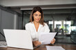 © Stock 4 You - Focused European Latin business woman accountant holding documents, work at laptop pc doing online data base trade market analysis. Portrait 40s mature hispanic businesswoman with paperwork in office