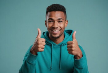 Wall Mural - A confident man with a bright smile and a teal hoodie gives two thumbs up against a teal backdrop.