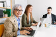 © Xavier Lorenzo - Smiling gray haired 60s senior businesswoman looking at camera sitting at workplace with young adult colleagues. Teamwork and business people concept.