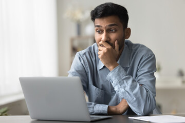 Bewildered confused or surprised Indian man sit at desk looking at laptop screen read news, experiences lack of understanding, feels confused, has problems with new apps or completing creative task