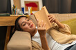 © Chanakon - Asian beautiful woman lay down on cozey couch sofa reading book in living room with warm light at night. People female stay home part time relax at home.