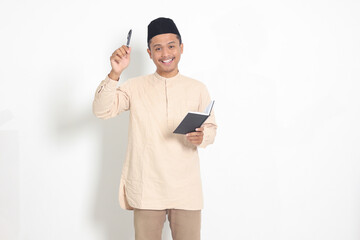 Wall Mural - Portrait of attractive Asian muslim man in koko shirt with peci reading a book, telling that he has an idea while pointing finger and pen. Isolated image on white background