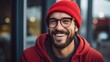 A close-up of a handsome smiling charismatic bearded man wearing a red beanie hat, hoodie and glasses looks away on the street. A creative happy person.