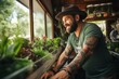 A bearded smiling man with tattoos is engaged in growing fresh herbs at home by the window