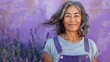 A joyful woman with gray hair wearing a blue denim apron standing in front of a purple wall with lavender flowers smiling warmly.