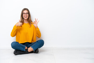 Wall Mural - Young caucasian woman sitting on the floor isolated on white background showing ok sign and thumb up gesture